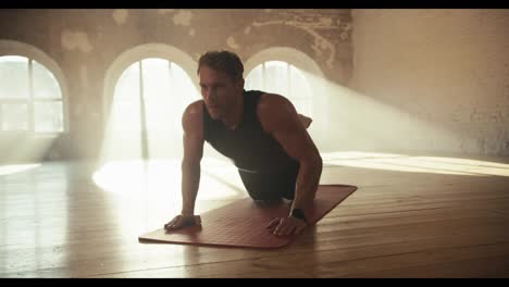 A-male-athlete-in-a-black-athletic-tight-fitting-uniform-does-push-ups-on-his-knees-on-a-special-mat-in-a-sunny-brick-hall