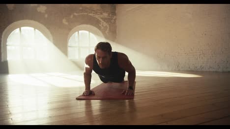 A-male-athlete-in-a-black-sports-summer-uniform-does-push-ups-on-a-special-mat-in-a-sunny-brick-hall-on-the-floor