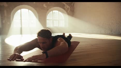 A-male-athlete-in-a-black-sportswear-summer-uniform-does-low-boat-exercises-in-a-brick-sunny-gym