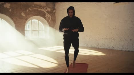A-man-in-a-black-hooded-sportswear-warms-up-and-runs-in-place-on-a-special-mat-in-a-sunny-brick-hall