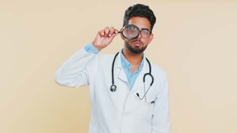 Indian-doctor-man-holds-magnifying-glass-near-face-looking-at-camera-with-big-zoomed-eye,-analyzing