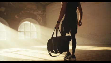 Athlete-man-in-black-sportswear-in-summer-uniform-brings-a-backpack-and-lays-out-his-fitness-equipment-in-a-sunny-brick-hall