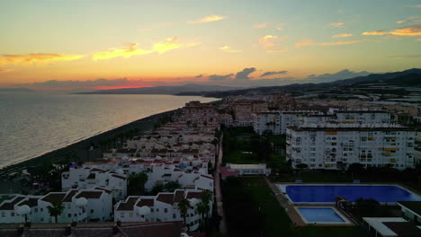 Aerial-dolly-backwards-shot-of-the-beautiful-idyllic-spanish-city-of-Malaga,-Costa-del-Sol-right-on-the-coast-in-front-of-the-calm-sea-with-historic-buildings-at-golden-hour
