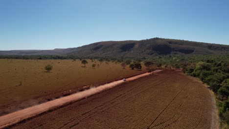 drone-view-of-a-moving-car-raising-dust-on-a-dirt-road-beauty-street-in-Chapada-dos-Veadeiros,-Goiás,-Brazil
