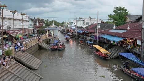 A-boat-coming-in-from-Mae-Klong-River-into-Amphawa-Floating-Market-Canal,-passing-by-rows-of-souvenir-shops,-restaurants-and-parked-wooden-boats-on-the-side-of-the-canal,-at-Samut-Songkhram,-Thailand