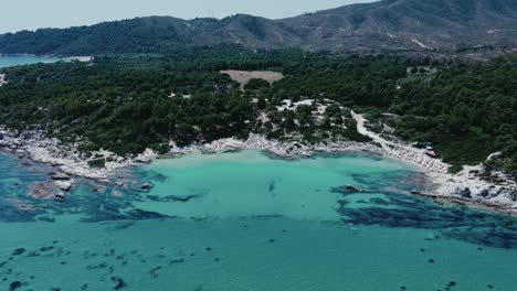 Aerial-establishing-shot-of-crystal-clear-water-with-many-tourist-swimming-in-bay-on-Chalkdiki,-Greece
