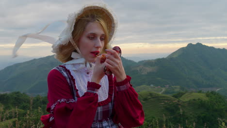 Blonde-woman-dressed-in-mountain-village-girl-outfit-applies-lipstick-on-windy-mountain-top