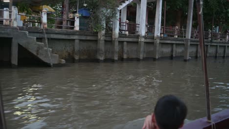 Ein-Kleiner-Junge-Fährt-Am-Ufer-Des-Schwimmenden-Marktkanals-Von-Amphawa-Entlang-Und-Betrachtet-Seine-Umgebung,-Während-Das-Boot-In-Richtung-Mae-Klong-Fluss-In-Der-Provinz-Samut-Songkhram-In-Thailand-Fährt