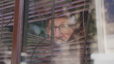 Old-man-in-glasses-opens-blinds-and-looks-out-window,-from-outside