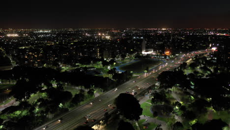 Eine-Luftaufnahme-Nach-Einbruch-Der-Dunkelheit,-Die-Eine-Belebte-Autobahn-Und-Einen-Park-In-Buenos-Aires-Zeigt,-Wo-Der-Verkehr-Zwischen-Den-Beleuchteten-Gebäuden-Der-Stadt-Fließt