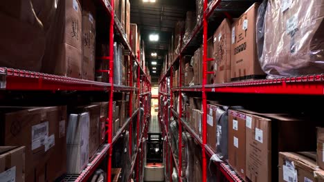 A-Young-Man-Drives-Leaving-an-Industrial-Warehouse-Lift-Pick-Truck-Forklift-Under-a-falling-Drone-Camera-Flying-Between-Two-Racks-of-Merchandise-Products-Inside-a-Shipping-Distribution-Centre