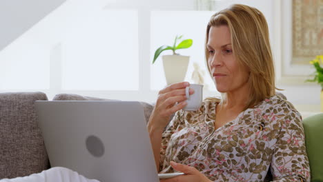 Woman-having-coffee-while-using-laptop-on-sofa-4k