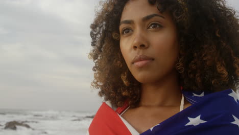 Vorderansicht-Einer-Afroamerikanischen-Frau,-Die-In-Eine-Amerikanische-Flagge-Gehüllt-Ist-Und-Am-Strand-Steht-4k