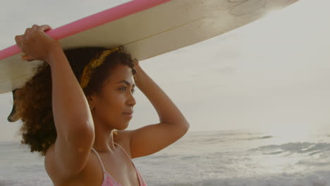 Side-view-of-African-American-female-surfer-carrying-surfboard-on-her-head-on-the-beach-4k