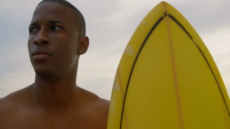 Front-view-of-African-American-male-surfer-standing-with-surfboard-on-the-beach-4k