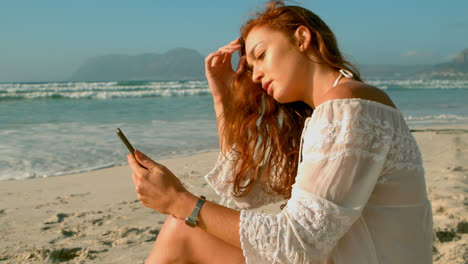 Caucasian-woman-using-mobile-phone-while-sitting-on-beach-in-the-sunshine-4k