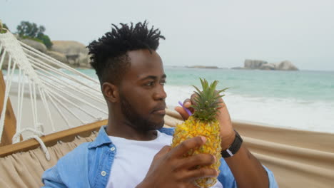 Front-view-of-African-american-man-drinking-pineapple-juice-on-the-beach-4k