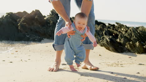 Front-view-of-mid-adult-caucasian-father-helping-baby-to-walk-at-beach-on-a-sunny-day-4k