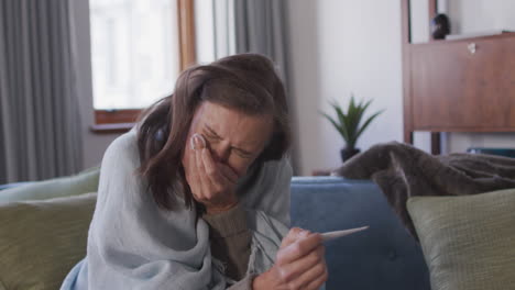 Woman-holding-thermometer-sneezing-while-sitting-on-couch-at-home