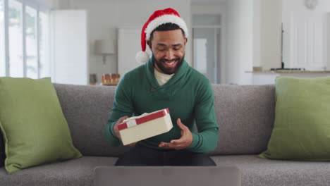 Man-wearing-Santa-hat-opening-gift-box-while-having-video-chat-on-his-laptop