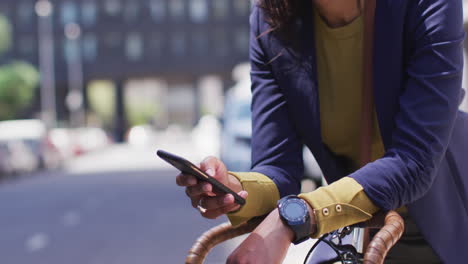 African-american-woman-using-smartphone-in-street-leaning-on-bicycle