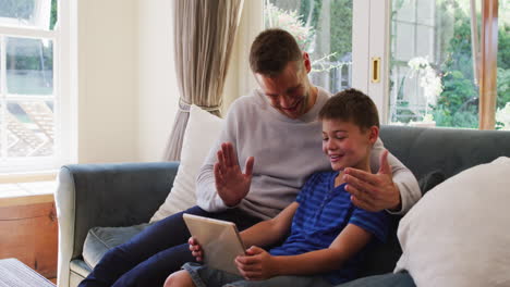Caucasian-man-and-son-smiling-while-having-a-video-call-on-digital-tablet-sitting-on-the-couch-at-ho