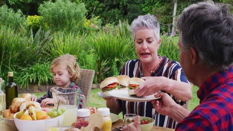 Smiling-caucasian-senior-woman-serving-family-having-celebration-meal-together-in-garden