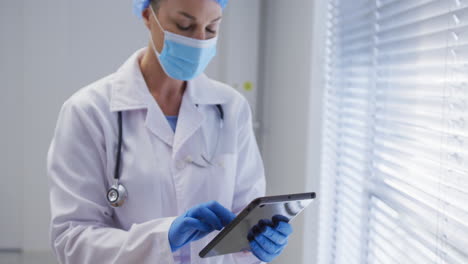 Caucasian-female-doctor-wearing-face-mask-and-surgical-gloves-using-tablet