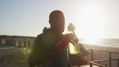 African-american-man-pouring-water-in-his-head,-taking-break-in-exercise-outdoors-by-the-sea