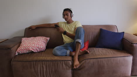 Mixed-race-man-wearing-headphones-sitting-on-sofa-using-smartphone-and-smiling