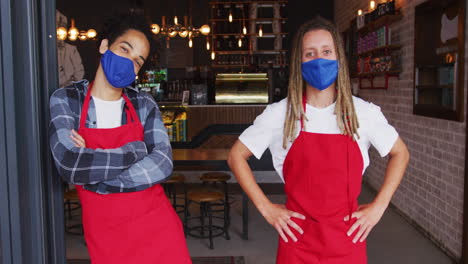 Two-diverse-male-baristas-wearing-face-masks-and-aprons-standing-in-doorway-of-cafe
