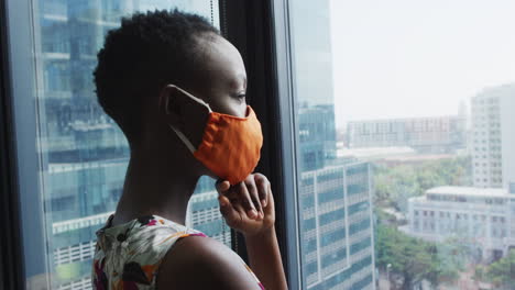 Thoughtful-african-american-woman-wearing-face-mask-looking-out-of-window-at-modern-office
