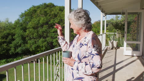 Caucasian-senior-woman-holding-coffee-cup-waving-while-standing-in-the-balcony-at-home