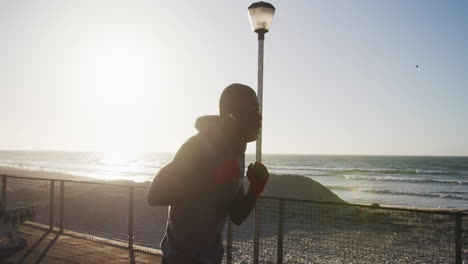 Fokussierter-Afroamerikanischer-Mann,-Der-Boxt-Und-Läuft-Und-Draußen-Am-Meer-Trainiert