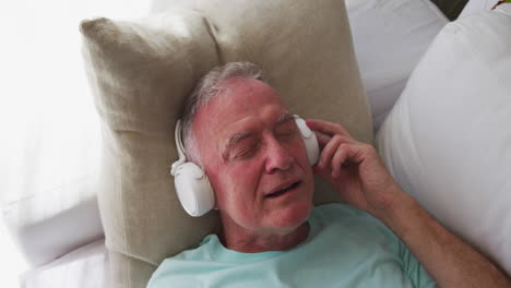 Kaukasischer-älterer-Mann-Mit-Kopfhörern,-Der-Singt,-Während-Er-Zu-Hause-Auf-Der-Couch-Liegt