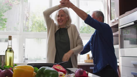 Glückliches-älteres-Kaukasisches-Paar-Zu-Hause,-Das-In-Der-Küche-Tanzt-Und-Lächelt,-Während-Es-Eine-Mahlzeit-Zubereitet