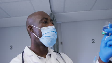 Afroamerikanischer-Männlicher-Arzt-Mit-Gesichtsmaske,-Der-Die-Spritze-Im-Krankenhaus-Mit-Covid-19-Impfstoff-Füllt