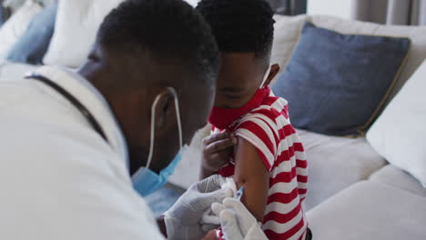 Afroamerikanischer-Arzt-Mit-Gesichtsmaske-Injiziert-Einem-Jungen-Zu-Hause-Den-Covid-19-Impfstoff
