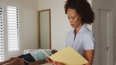 Portrait-of-mixed-race-female-physiotherapist-holding-notebook-and-writing-looking-at-camera