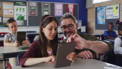 Diverse-male-teacher-helping-a-schoolgirl-sitting-in-classroom-using-tablet