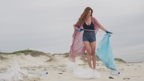 Caucasian-woman-wearing-latex-gloves-collecting-rubbish-from-the-beach-using-grabber-stick