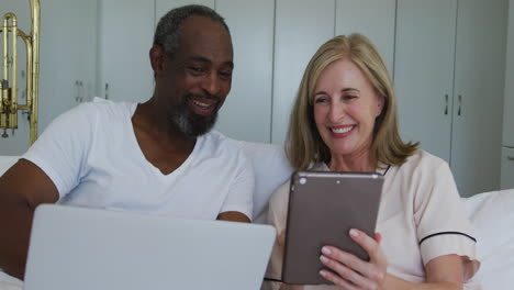 Diverse-senior-couple-sitting-in-bed-using-laptop-and-tablet-talking-and-lsmiling