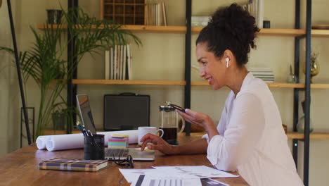 Mixed-race-woman-using-laptop-and-talking-on-smartphone-while-working-from-home