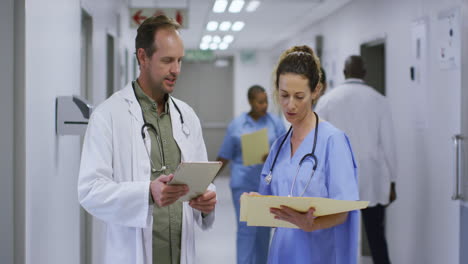 Caucasian-male-and-female-doctor-talking-in-hospital-corridor-looking-at-tablet-and-patient-files