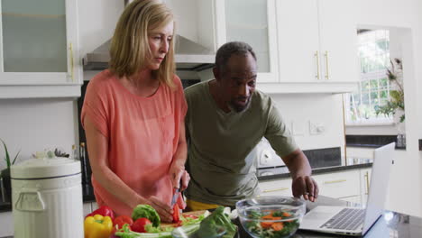 Diverse-senior-couple-preparing-food-in-kitchen-following-recipe-on-laptop