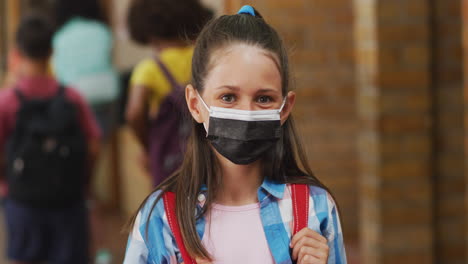 Portrait-of-caucasian-schoolgirl-wearing-face-mask,-standing-in-corridor-looking-at-camera
