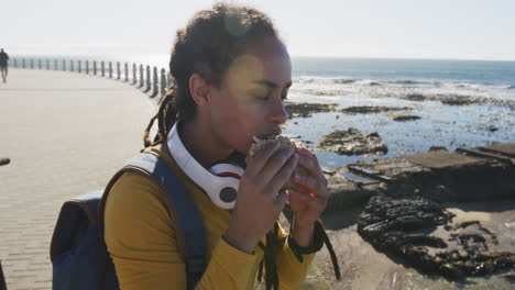 African-american-woman-sitting-eating-sandwich-promenade-by-the-sea
