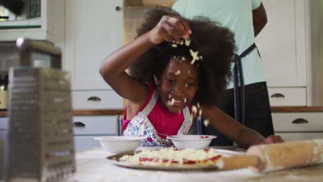 Hija-Afroamericana-Haciendo-Pizza-En-La-Cocina,-Su-Padre-Cocinando-En-Segundo-Plano.