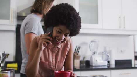 African-american-woman-holding-coffee-cup-talking-on-smartphone-in-the-kitchen-at-home