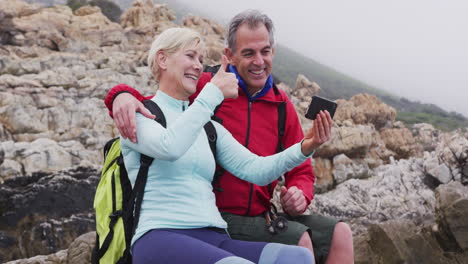 Senior-hiker-couple-with-backpacks-sitting-on-the-rocks-and-waving-while-having-a-video-call